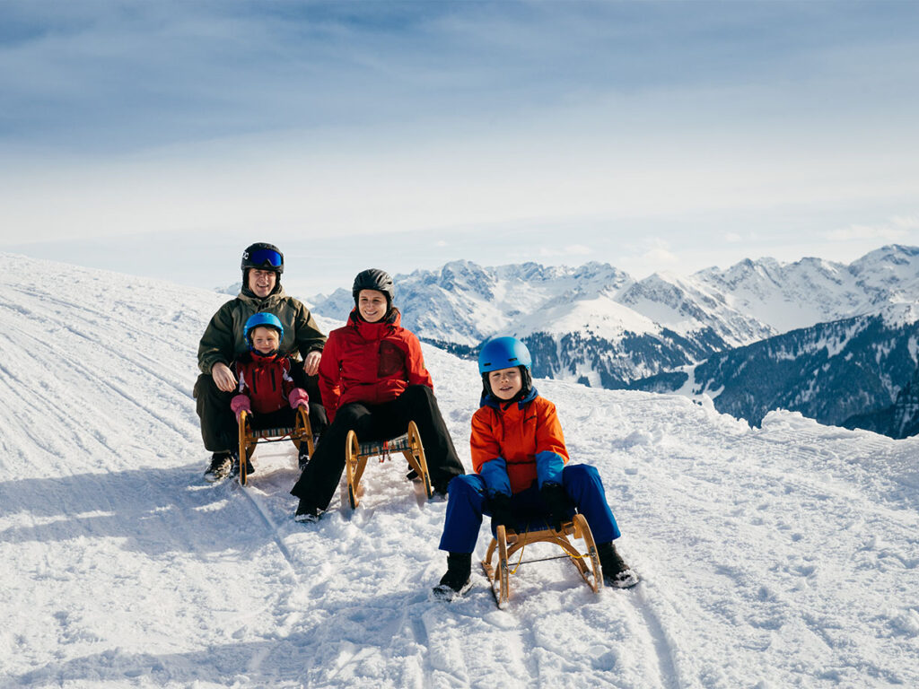 Familie auf drei Schlitten auf Rodelbahn in Bezau mit Bergen im Hintergrund